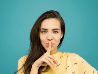 portrait photo of woman in yellow t shirt doing the shh sign while standing in front of blue background